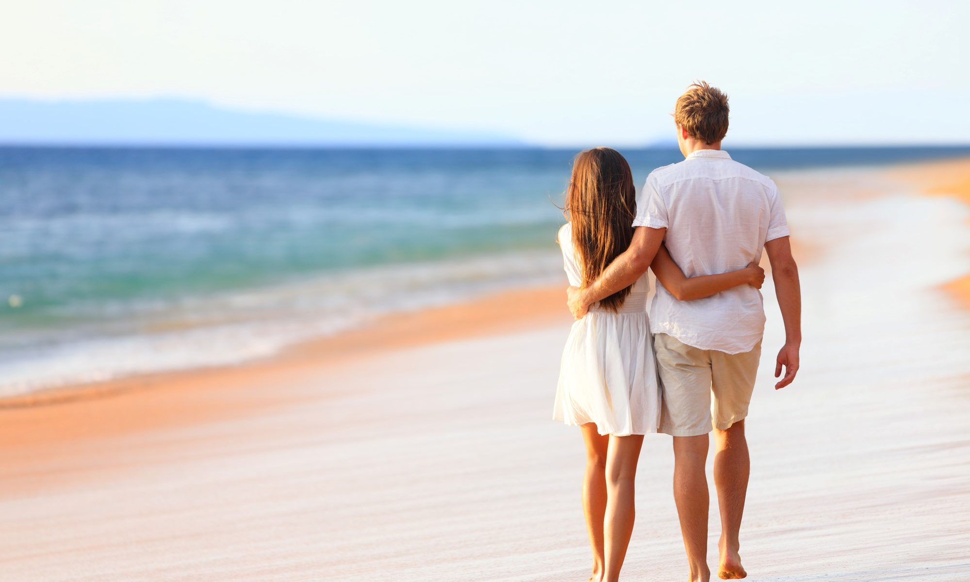 A couple walking hand in hand on a beach at sunset with the silhouette of the Bitcoin logo in the ocean.