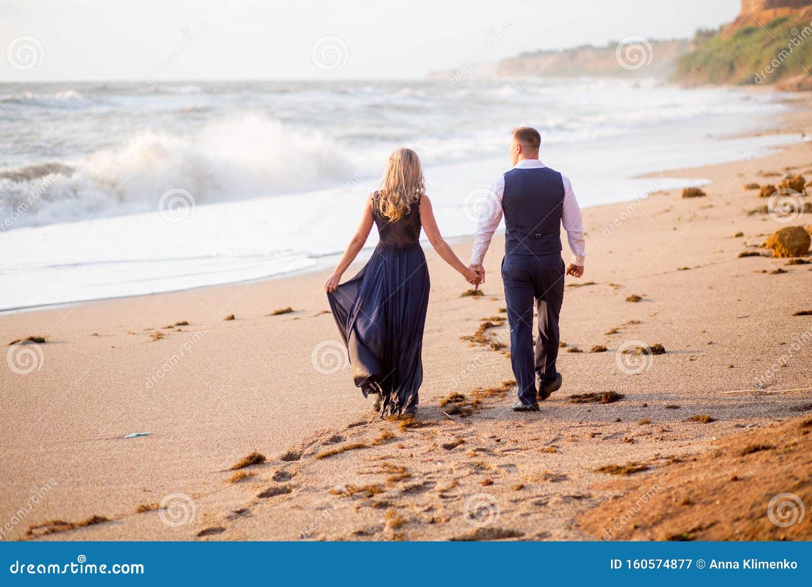 A couple walking hand in hand on a beach during sunset, with colorful sky and gentle waves.