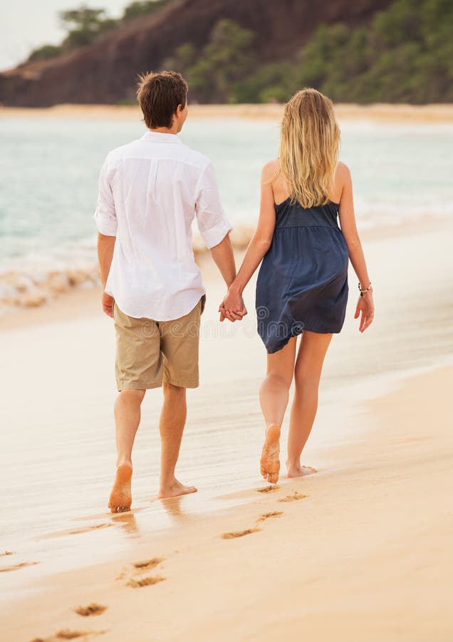 A romantic couple walking hand-in-hand on a beach during sunset.